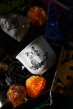 Skeleton couple teacup nestled in the Ancestor Alchemy Tea Kit with orange marigolds and a skull-shaped bottle.
