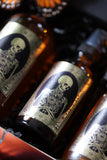 Close-up of amber Ancestor Alchemy bottles with gold skeleton-and-flower labels nestled in black shredded paper.