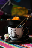 Skeleton couple teacup with a gold infuser on a black saucer beside a miniature sugar skull on a colorful striped textile.